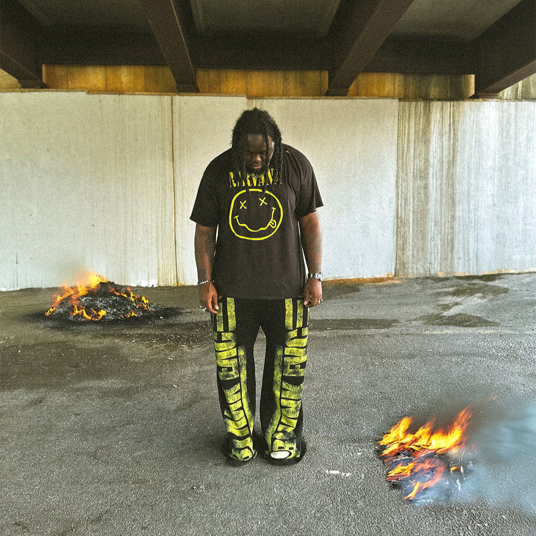 Person wearing a black t-shirt with a smiley face design and yellow pants standing between two fires under an overpass.