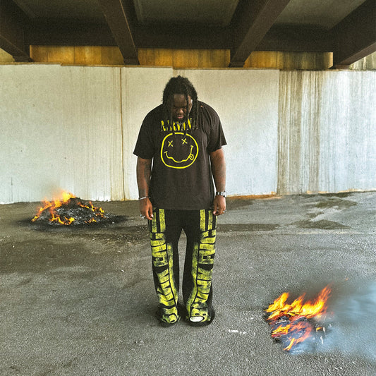 Person wearing a black t-shirt with a smiley face design and yellow pants standing between two fires under an overpass.